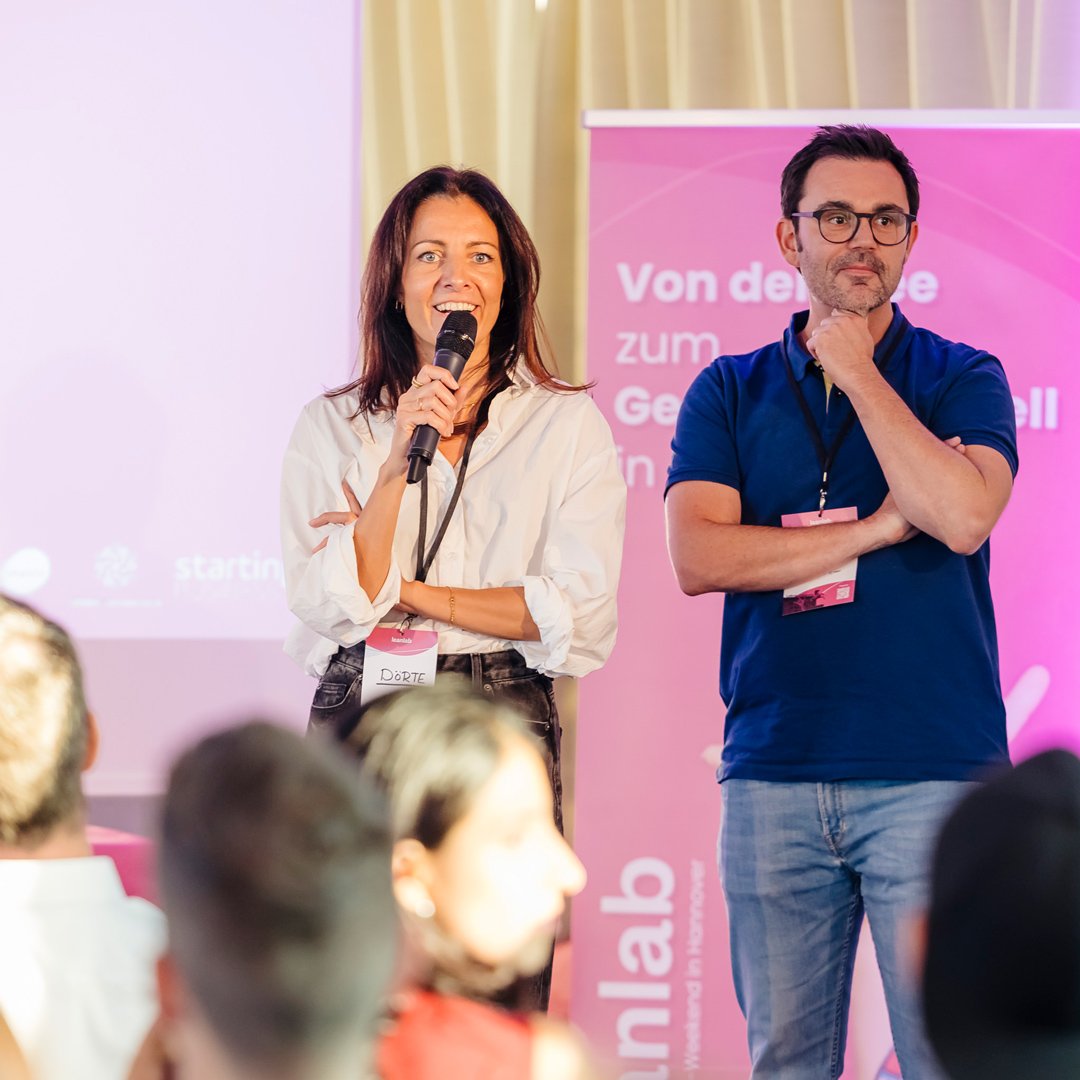 Woman holding a microphone and speaking at an event, standing next to a man with glasses and a beard, who has his hand on his chin. They are on stage with a pink banner in the background.