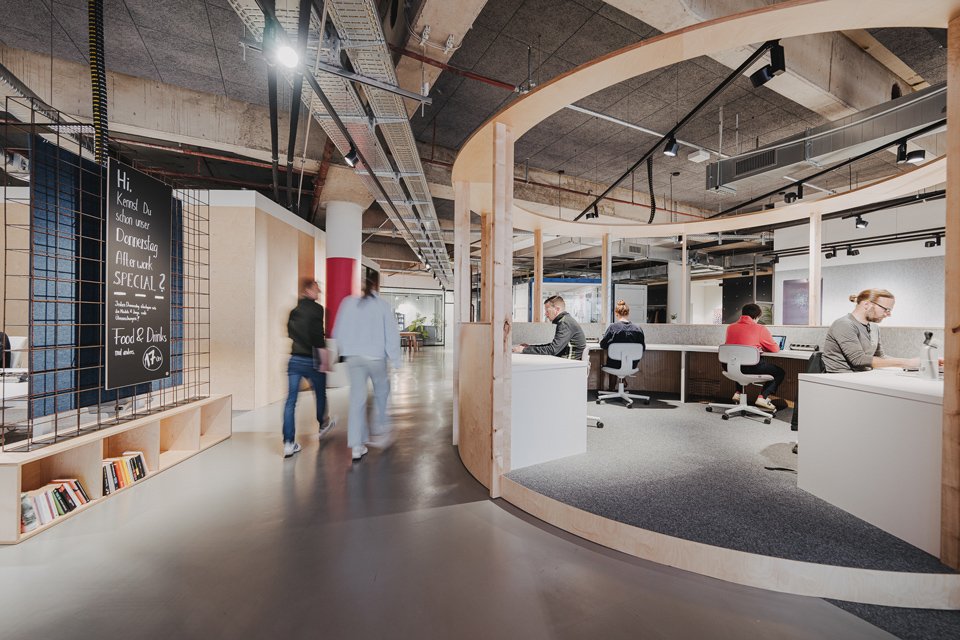 Open office space with curved wooden partition, people working at desks, and a chalkboard with a daily schedule.