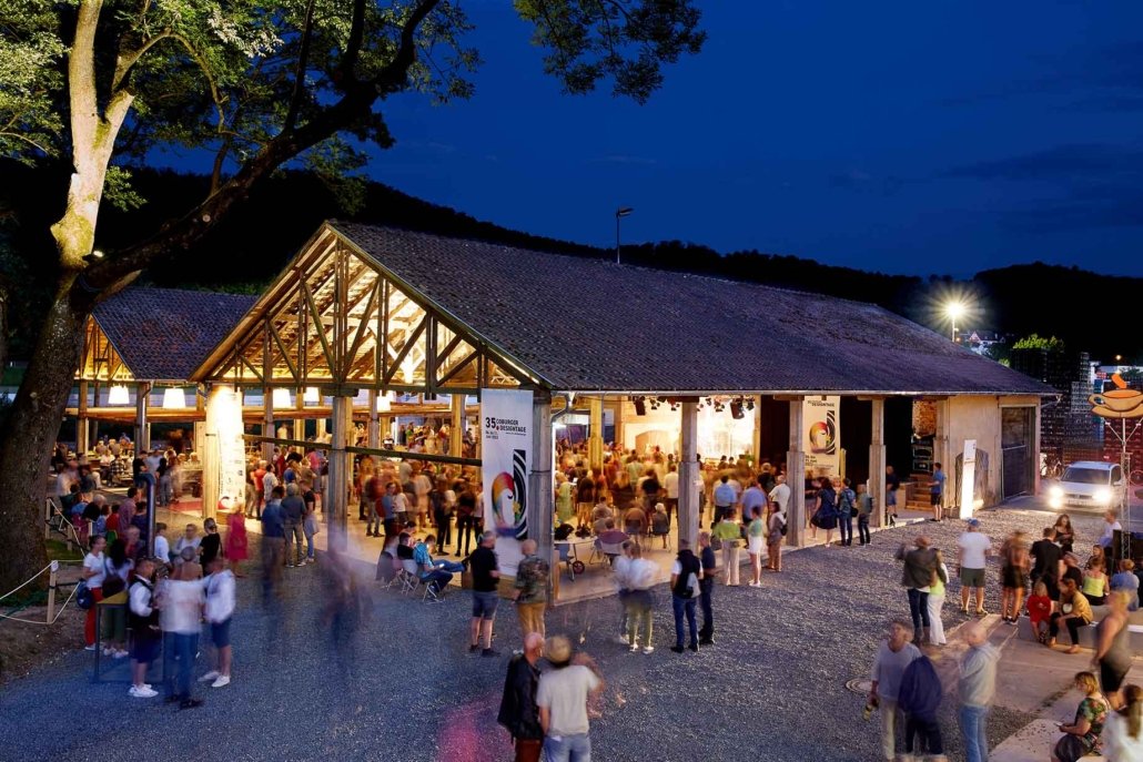 Outdoor event at night inside a large wooden pavilion with many people gathering, some sitting and some standing, under a tree, with a mountain backdrop and lights in the distance.