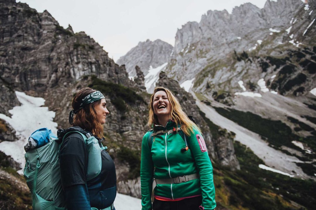 Two women with backpacks hiking in a mountainous area with rocky peaks and patches of snow, smiling and laughing.